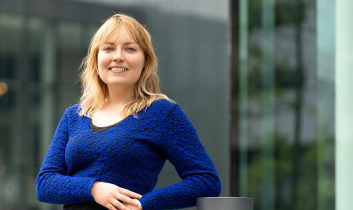 Woman in blue top smiling at camera in front of Alliance Manchester Business School building